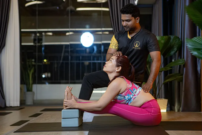A teacher supporting a student in a seated forward bend using a block